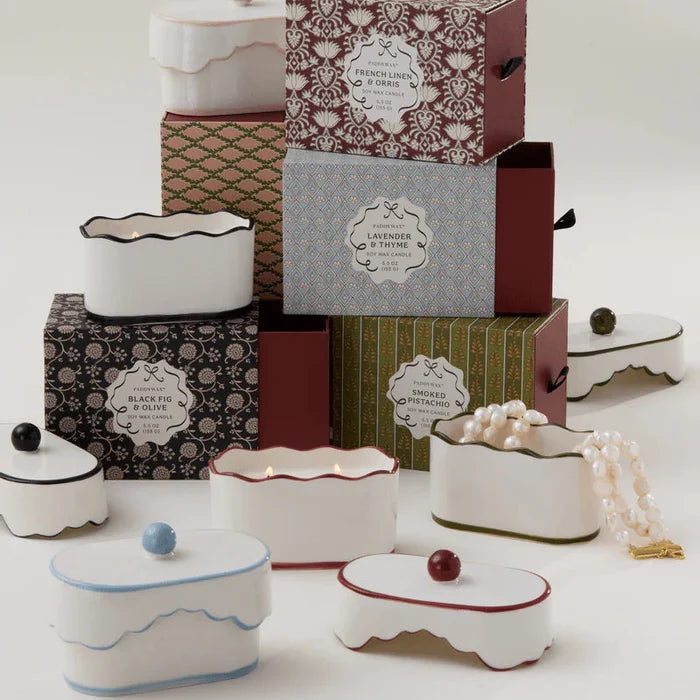 The 5.5oz Ceramic Dish Candle by Paddywax in white lidded containers is displayed with boxes labeled lavender, thyme, and black fig; a pearl necklace rests nearby.