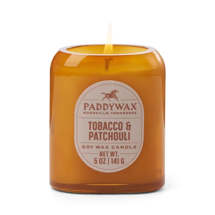 A lit 5oz Vista Candle in brown glass displays its luxurious tobacco & patchouli fragrance on a white background.