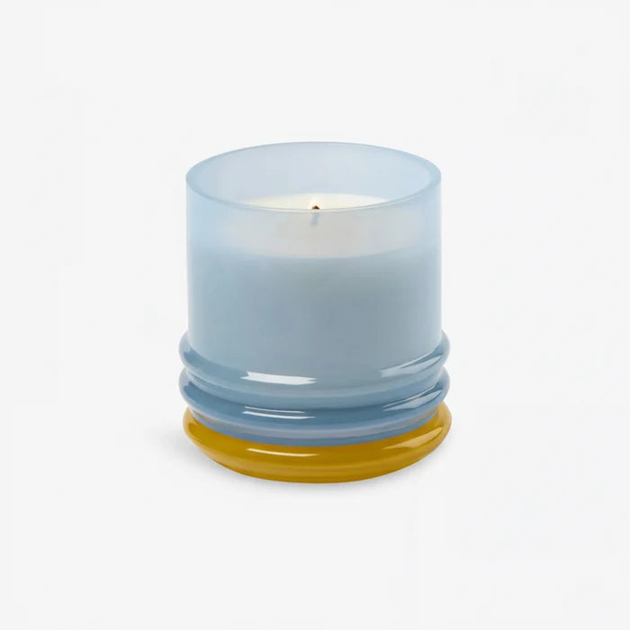 The 7oz Stacked Glass Candle, made with white soy wax, glows in a translucent, multi-colored glass holder with a rounded base featuring yellow and blue accents, set against a plain white background.