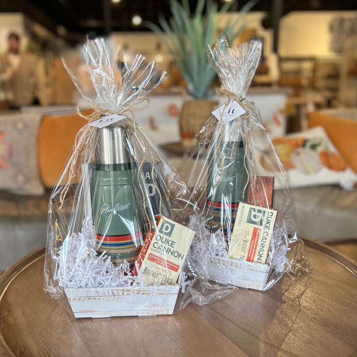Two Custom Gift Boxes from Lit Decor and Gift sit on a round wooden table. Each box includes a green thermos, Duke Cannon soap bars, shredded white paper, and a gift tag—everything for the perfect present in a cozy space.