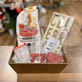 Create a Custom Gift Box from Lit Decor and Gift with a gold basket, mimosa muffin and bread mix, “Cheers” napkin set, “Clubber Mood” candle, matchbox, and two sprinkle tumblers—all arranged on a wooden surface.