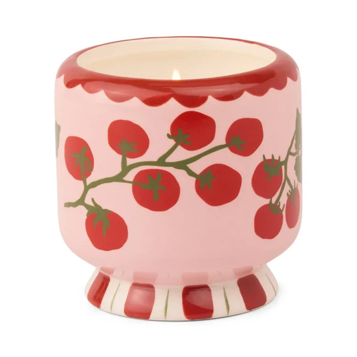 The A Dopo 8oz Ceramic Candle features a pink ceramic holder adorned with red tomatoes, green vines, a red scalloped rim, and a red-and-white striped base, holding a hand-painted candle inspired by Mediterranean fragrance.