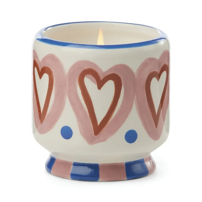 The A Dopo 8oz Ceramic Candle comes in a decorative ceramic holder with pink hearts, red outlines, blue dots, a blue and pink striped base, and is infused with a soothing Mediterranean fragrance.