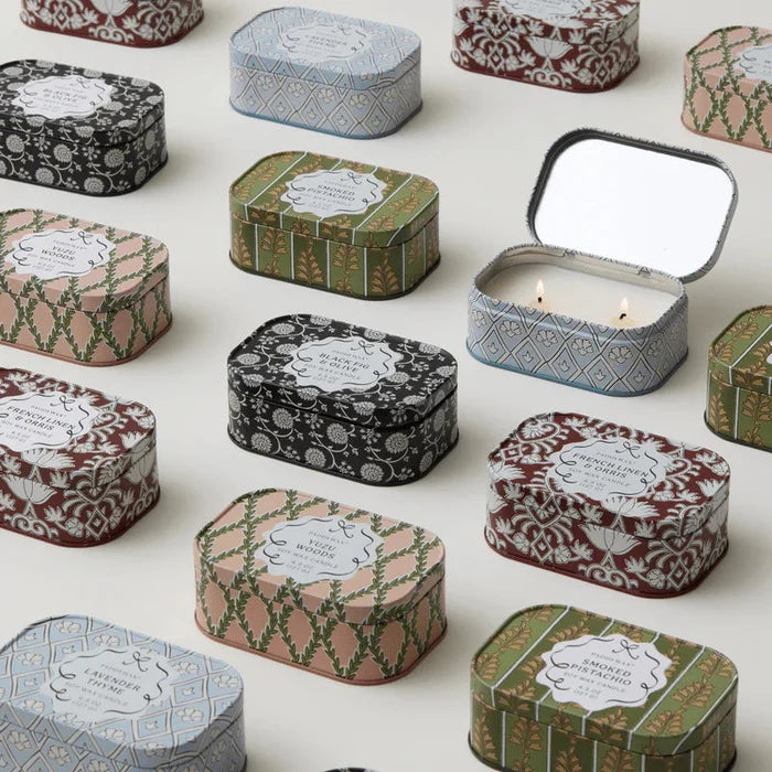 Several Cherie Tin 4.5 oz Candles featuring floral and geometric designs are arranged neatly on a light surface, with one tin open to reveal a lit double-wick decorative scented candle inside.