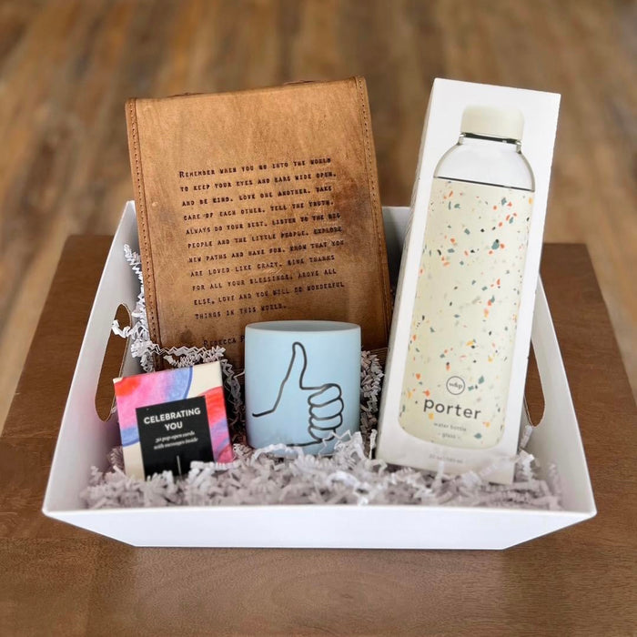 Create the perfect gift with Lit Decor and Gifts Custom Gift Boxes featuring a white tray, brown notebook, light blue thumbs-up mug, speckled Porter water bottle, and a vibrant CELEBRATING YOU box on white decorative paper.