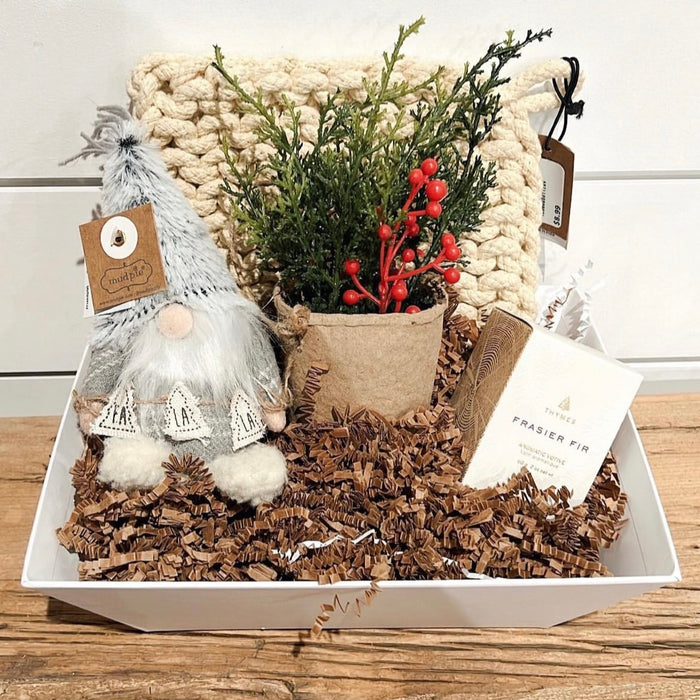 The Custom Gift Box by Lit Decor and Gift includes a knitted cream blanket, small plush gnome, faux plant with red berries, and Frasier Fir candle—artfully arranged on shredded brown paper in a white tray.