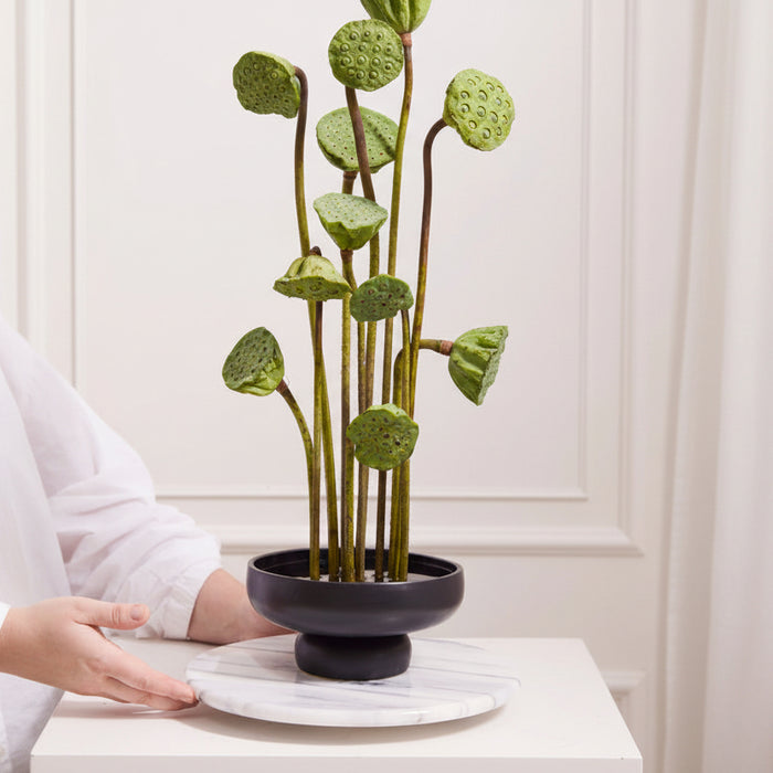 A person creates an ikebana arrangement with tall green lotus pods and stems in a glossy black compote on a round white marble tray, set against a light-colored wall.