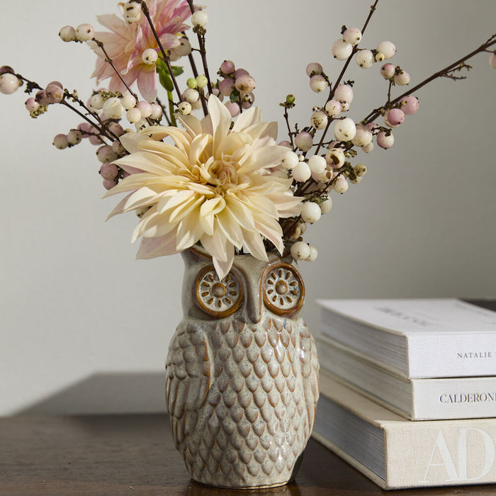 The Great Horned Owl Vase, featuring a grey reactive glaze, displays cream dahlias with white and pink berry sprigs on a wood surface next to stacked books.