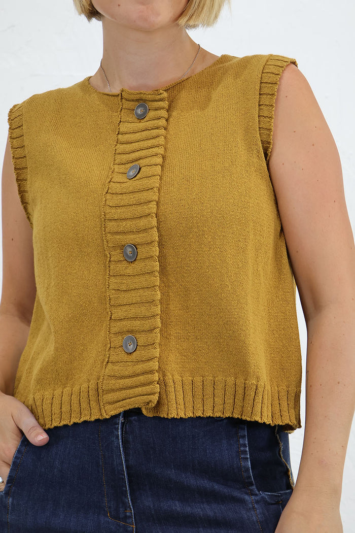 A sleeveless mustard yellow knit vest with a button-front closure and ribbed trim, shown on a model wearing blue jeans against a plain white background. Outfit by Lurdes Bergada at Blue