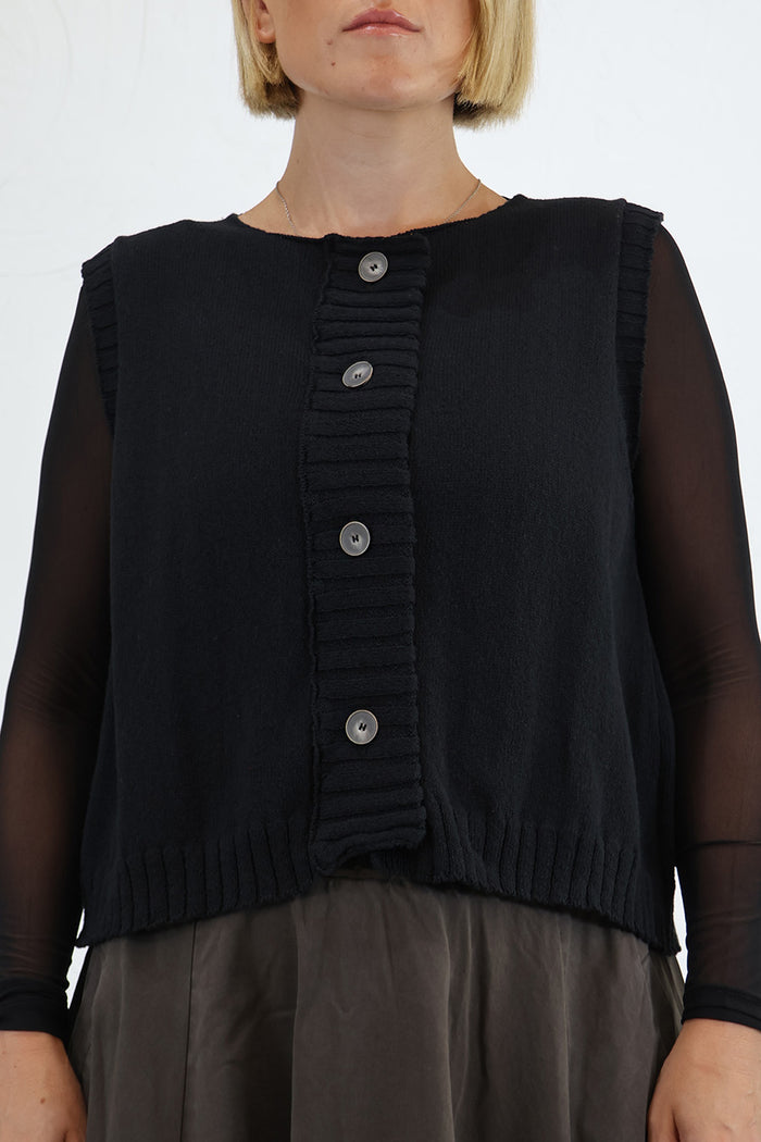 A close-up of a model wearing a black sleeveless ribbed knit vest with a button-front detail over sheer long-sleeve undergarments. Lurdes Bergada at Blue
