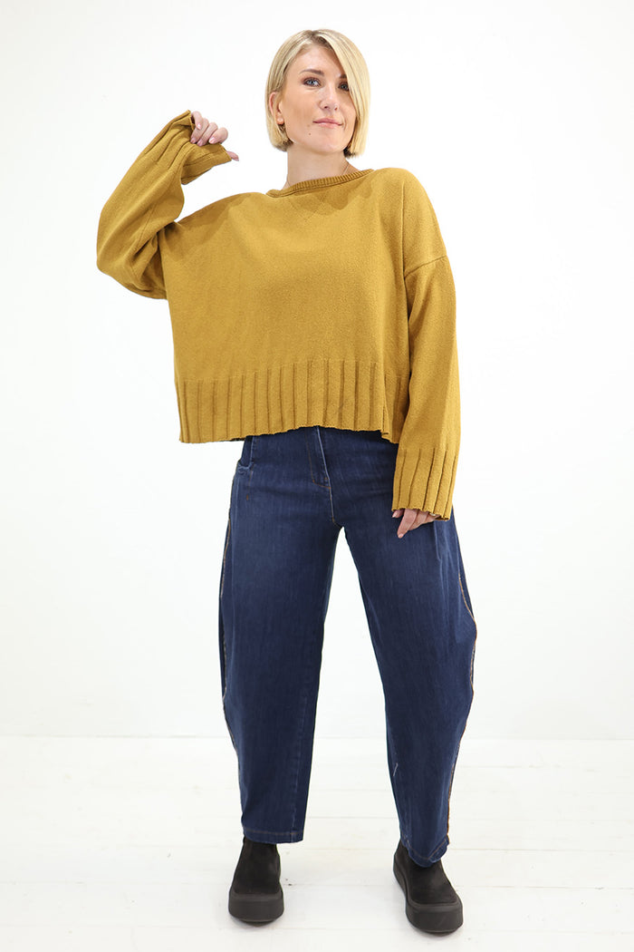 A woman modeling a cropped, oversized mustard yellow knit sweater with ribbed hem and cuffs, paired with dark blue baggy jeans and black slip-on shoes, against a plain white studio background. Lurdes Bergada at Blue