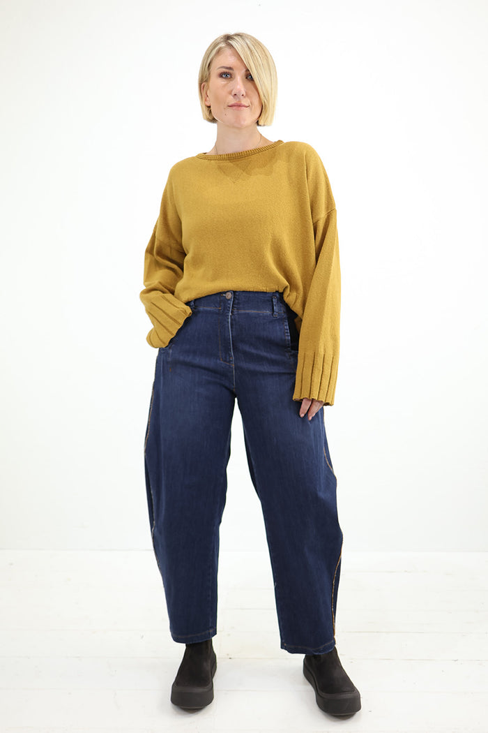 A woman modeling a mustard yellow knit sweater and dark blue wide-leg jeans against a clean white studio background. Outfit by Lurdes Bergada at Blue