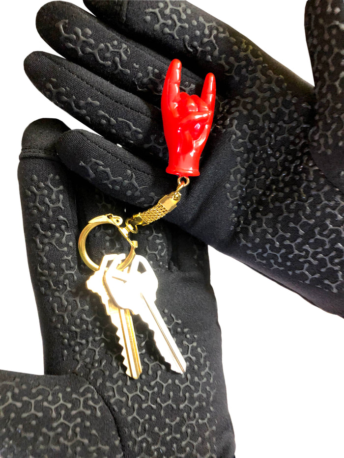 Red hand-shaped keychain making the “rock on” gesture, attached to a gold-colored chain and keyring on a hand. The glossy charm blends Italian Malocchio symbolism with rock culture, serving as a bold accessory for protection, style, or gifting.