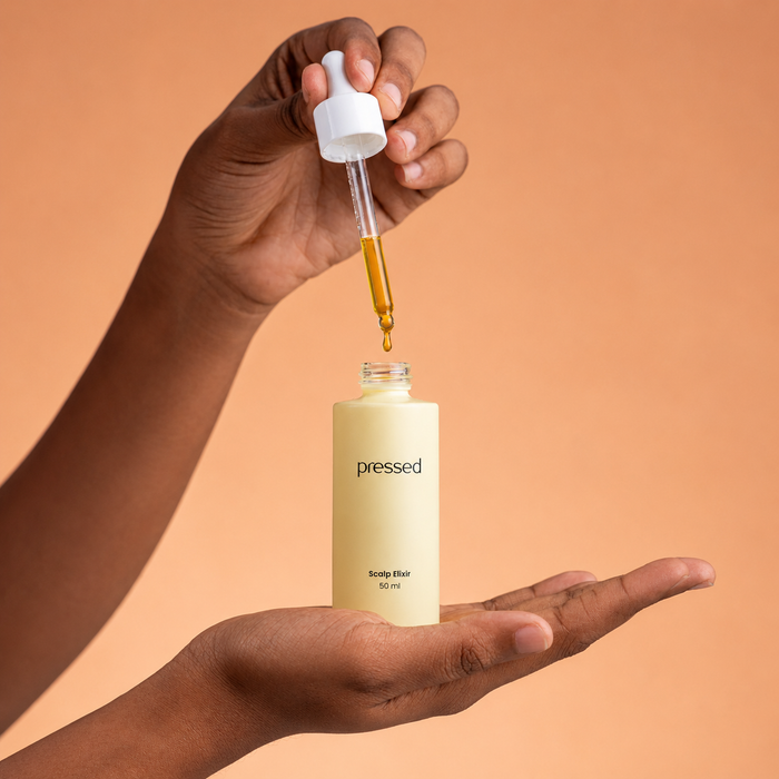 hand holding a pressed scalp elixir 50ml bottle while a dropper dispenses golden scalp oil above it against a warm beige background