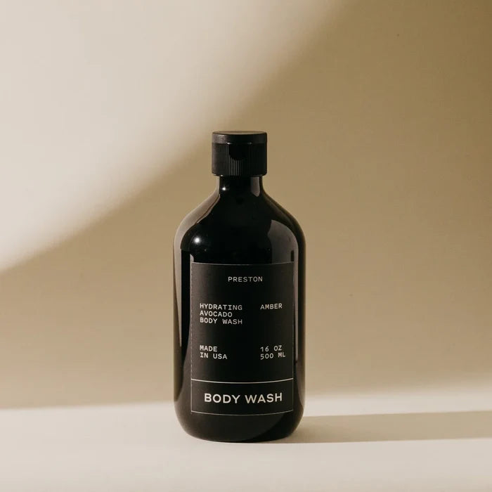 A dark bottle of Preston Body Wash stands on a light surface with a beige background. The label describes it as a moisturizing wash with hydrating avocado oil, made in the USA, and containing 16 oz (500 ml).