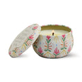A lit Printed Tulip Candle Tin sits open, revealing its scented glow. The round tin features a decorative pink, green, and yellow floral pattern; the matching lid rests nearby.
