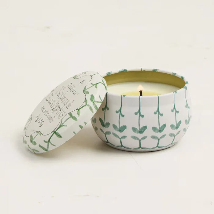 The Printed Tulip Candle Tin, featuring a white gardenia scent, sits lit in its round tin adorned with green leaf and bamboo patterns. The matching lid is set aside as the fresh, floral aroma fills the light-colored background.