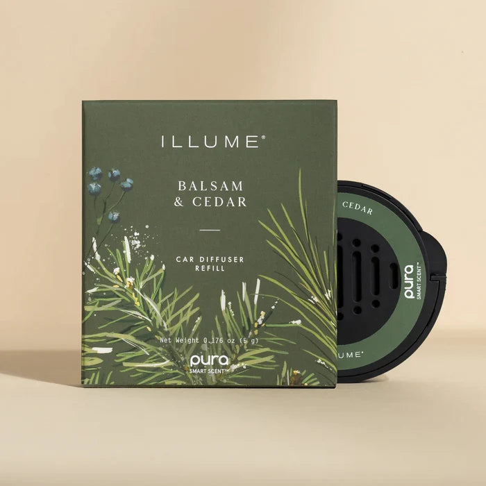 A round black Pura Car Diffuser Refill sits stylishly beside a green botanically illustrated box labeled “ILLUME Balsam & Cedar Car Diffuser Refill,” both displayed on a neutral beige background.