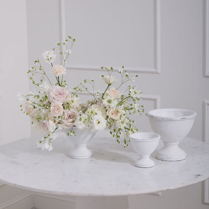 A modern white marble table features a floral vase with pale pink and white flowers, a Rustic White Compote, and two empty ceramic bowls of varying sizes, all set before a light paneled wall.