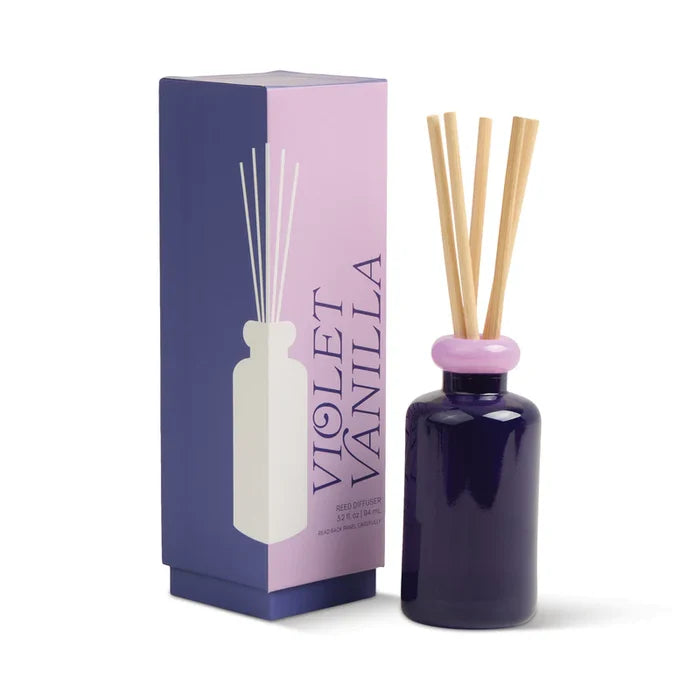 A violet Paddywax Stacked Reed Diffuser with a light pink cap and wooden reeds stands beside its matching Violet Vanilla box, which features bold purple text, a minimalist bottle illustration, and vibrant fragrance accents.