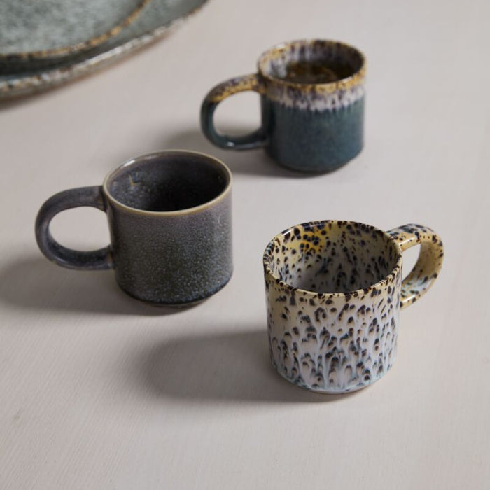 Three Starling Mug Collection stoneware mugs, featuring rounded handles and unique reactive glaze in gray, blue, and cream tones, are arranged on a light-colored surface.