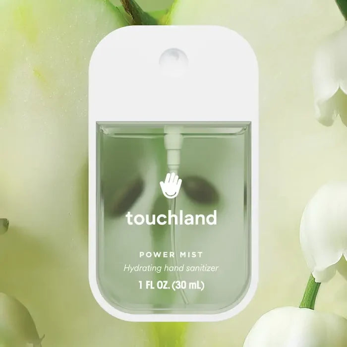 A rectangular Touchland Hand Sanitizer Mist bottle with clear green liquid and a white cap is displayed on a soft green background with white flowers, offering hydrated, fresh, and delicately scented hands.