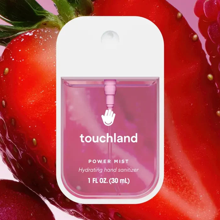 A pink bottle of Touchland Hand Sanitizer Mist is centered before a close-up of fresh strawberries. Its delicately scented formula hydrates hands, and the sleek container holds 1 fl oz (30 mL).