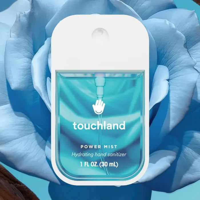 A bottle of Touchland Hand Sanitizer Mist, in clear blue with white text and a modern design, is displayed before a large blue rose—perfect for keeping hands fresh and delicately scented.