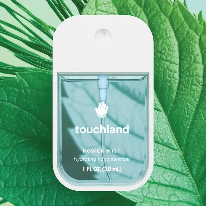 A clear rectangular Touchland Hand Sanitizer Mist bottle stands before overlapping green leaves, its logo and 1 fl oz (30 mL) label visible—ideal for hydrated hands with a delicately scented finish.