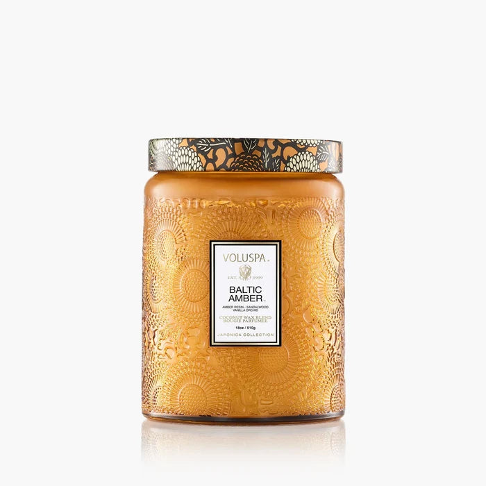 The Voluspa 18oz Glass Jar Candle features amber-colored embossed glass with floral patterns and a metallic lid, labeled "Voluspa Baltic Amber." Made from coconut wax, the candle is displayed against a white background.
